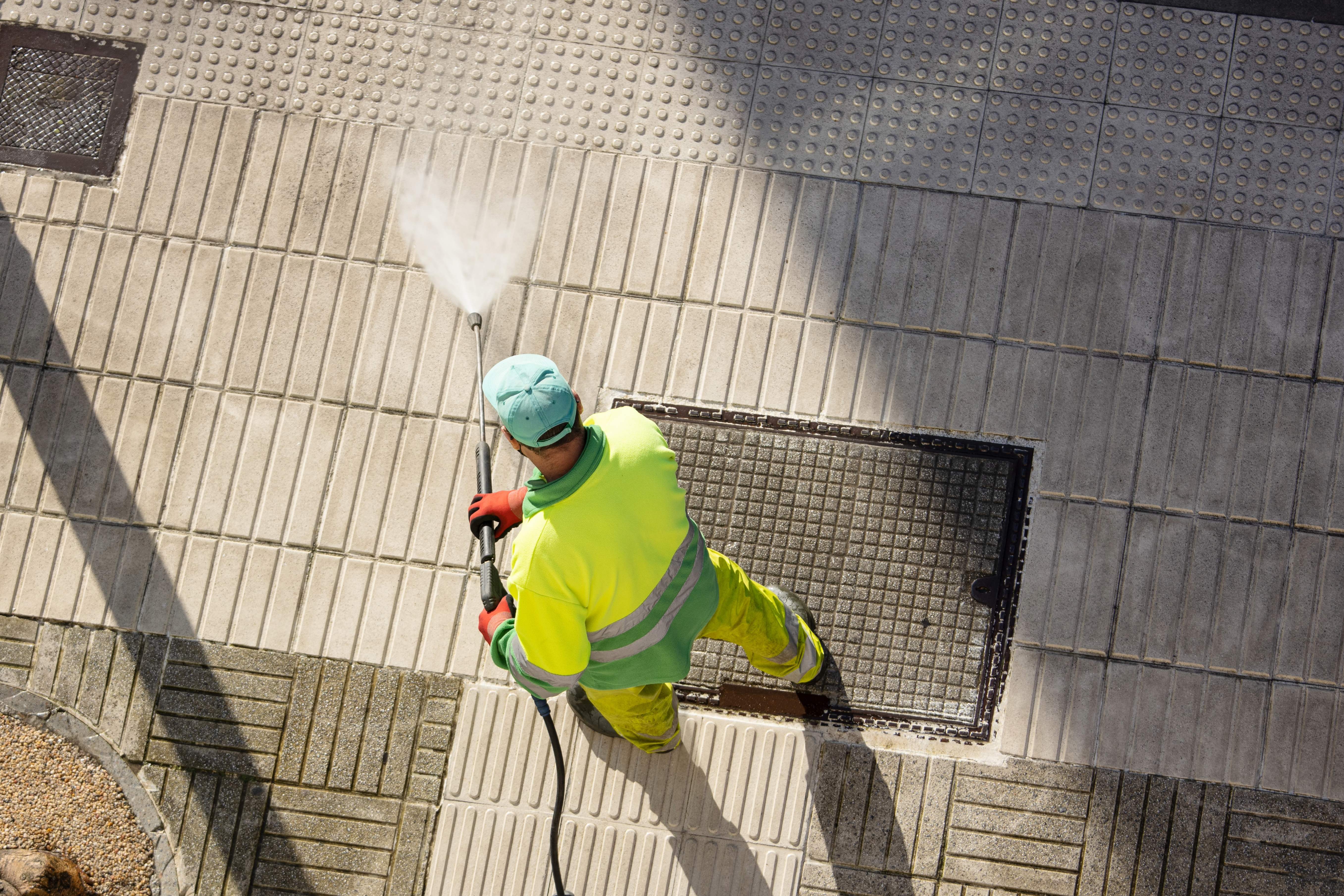 Aerial overhead view of professional worker in high-visibility yellow safety jacket and green cap using pressure washing equipment on various patio surfaces including concrete pavers